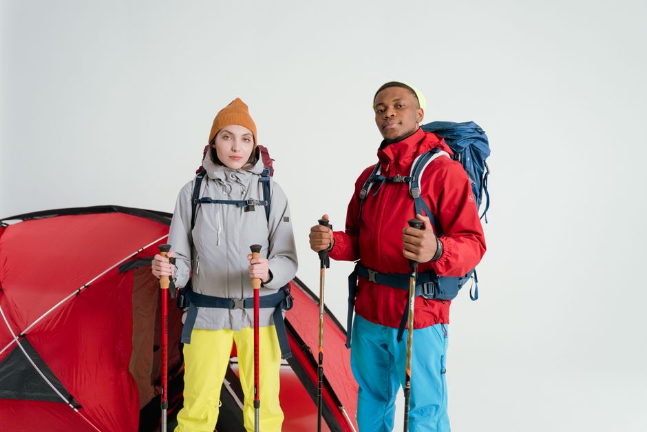 Two adults equipped with camping gear and hiking sticks pose in front of a tent on a white background.