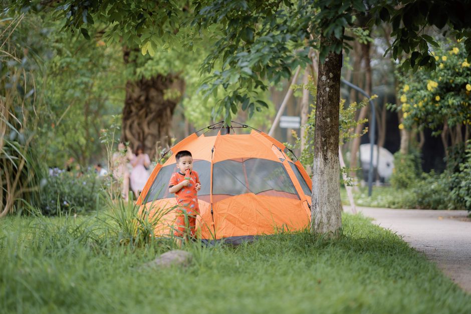 A young boy in an orange outfit stands beside a vibrant orange tent in a lush green park, enjoying a camping experience.