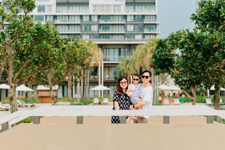 Positive young Asian couple with child wearing casual clothing and stylish sunglasses while standing among green tropical trees and looking at camera