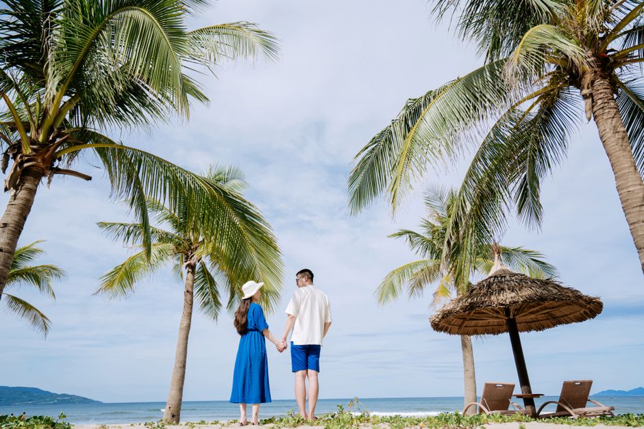 A couple holds hands under palm trees on a sunny tropical beach in summer.