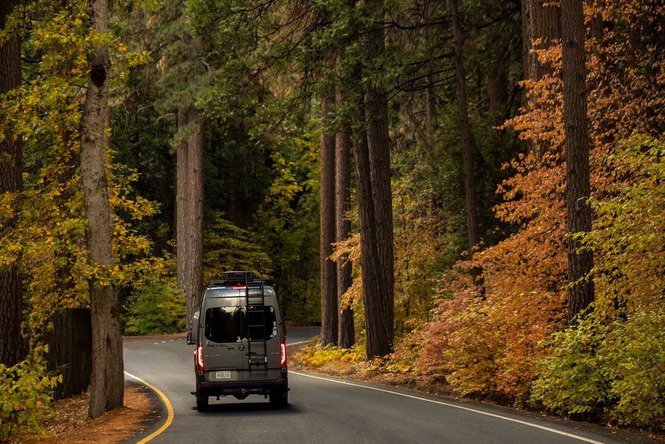 A camper van travels down a winding road surrounded by vibrant autumn foliage.