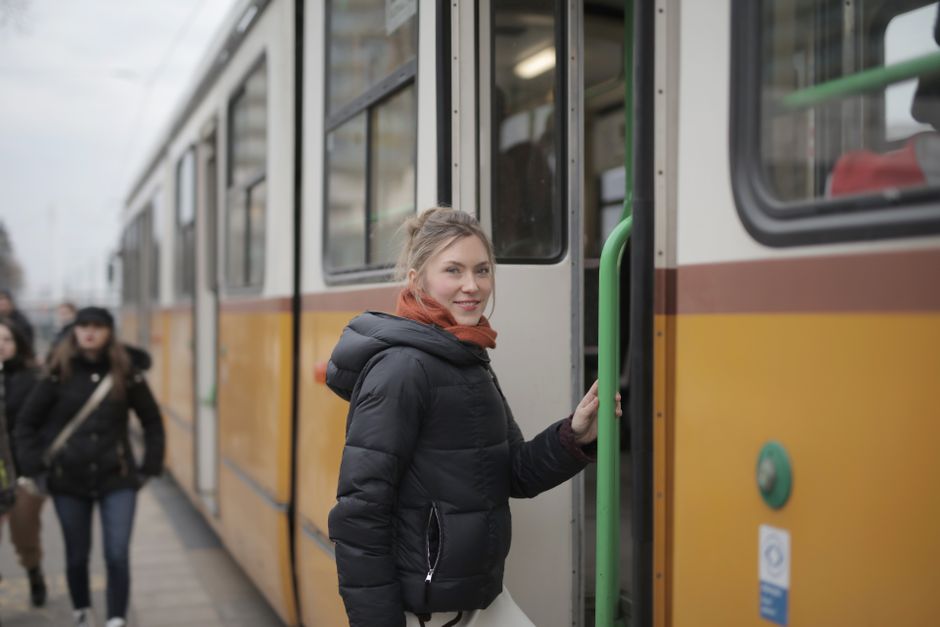Positive smiling female passenger in black warm jacket and scarf standing near opened tram door on urban street in cold season while entering inside and looking at camera