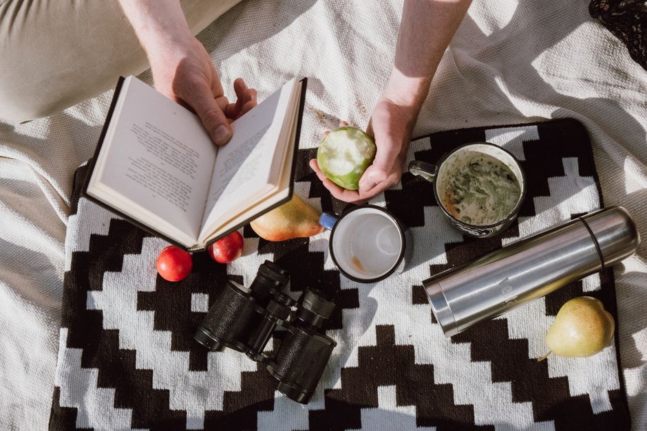 A serene picnic setting with a book, fruit, and binoculars on a patterned blanket.
