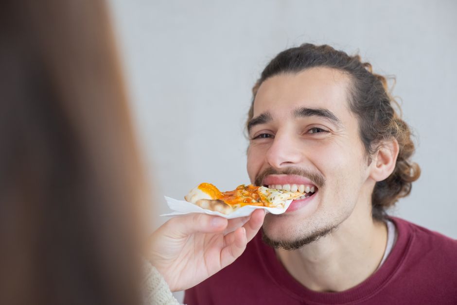 Smiling young man with facial hair enjoying a slice of pizza indoors.