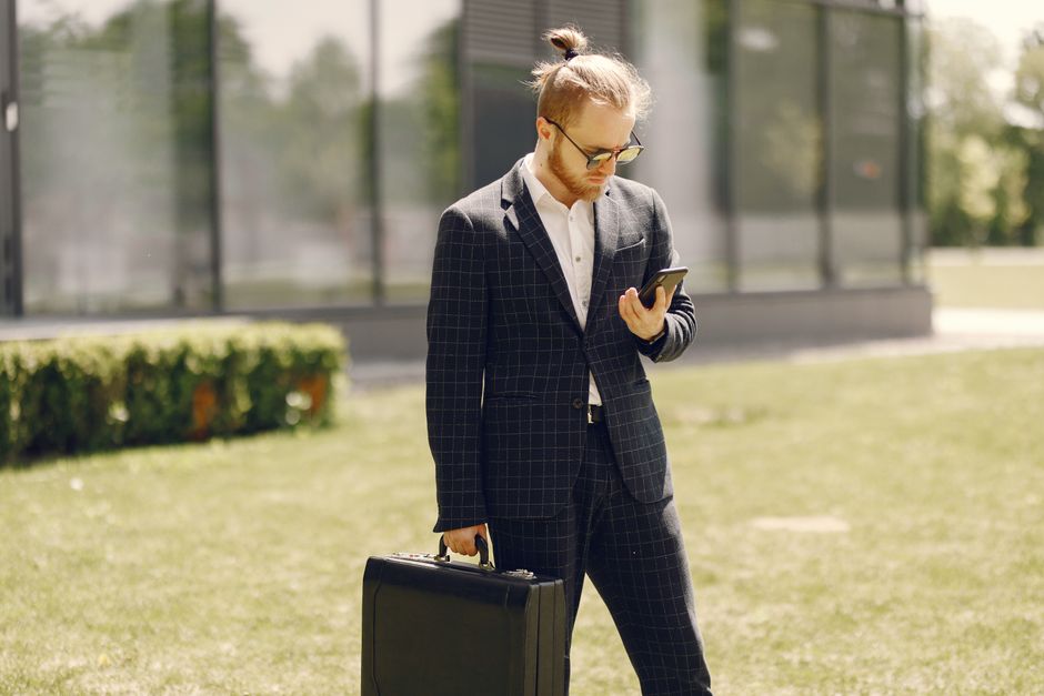 Young businessman in a suit checks his phone outside a modern building on a sunny day.