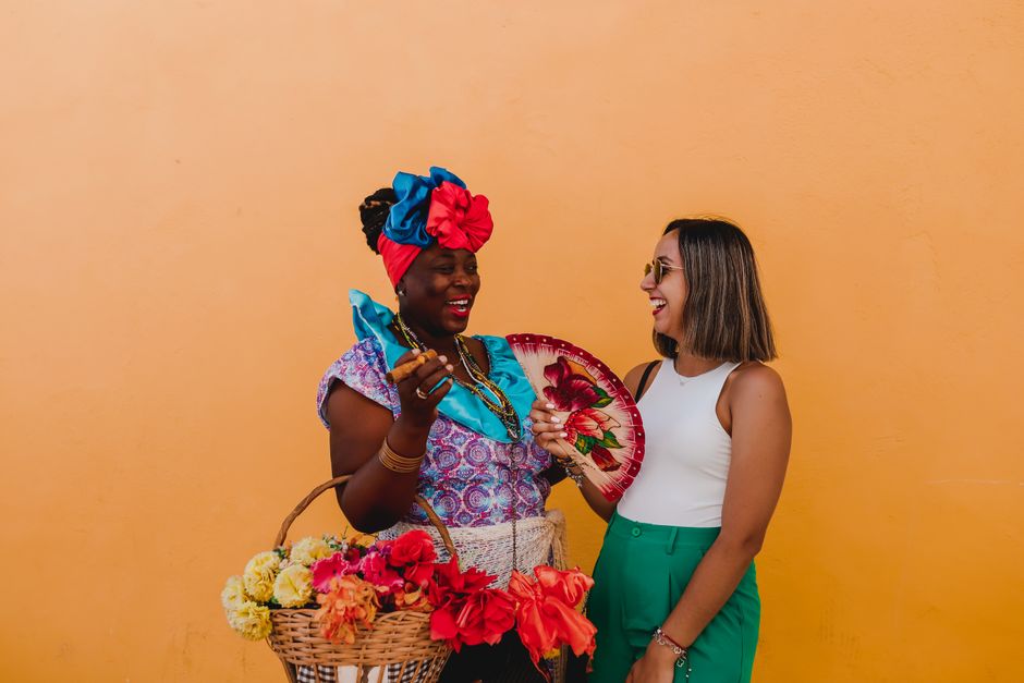 Two women laughing together while holding flowers and a fan against a bright orange background.