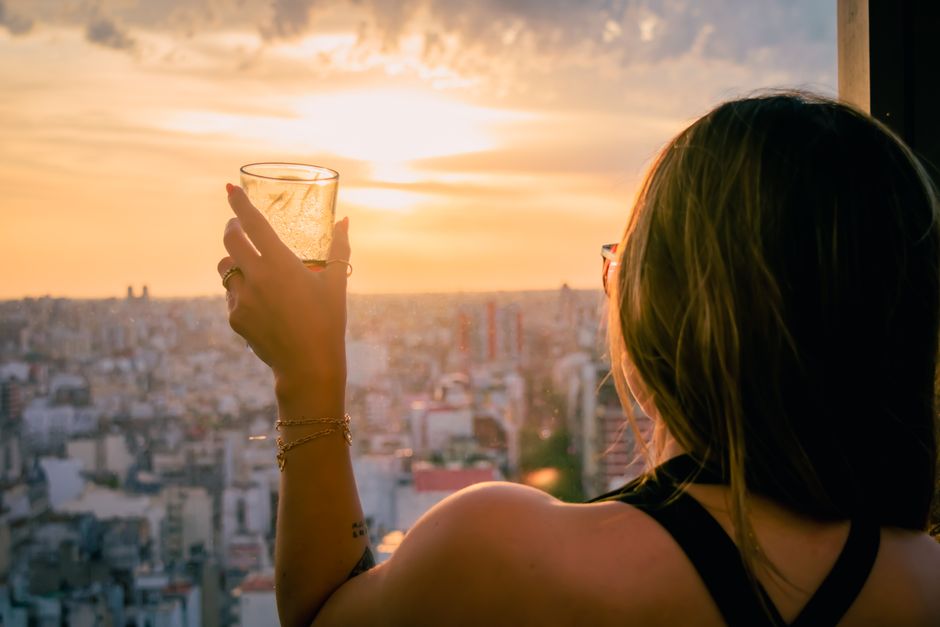 A woman holds a drink while overlooking a vibrant city skyline during a stunning sunset.