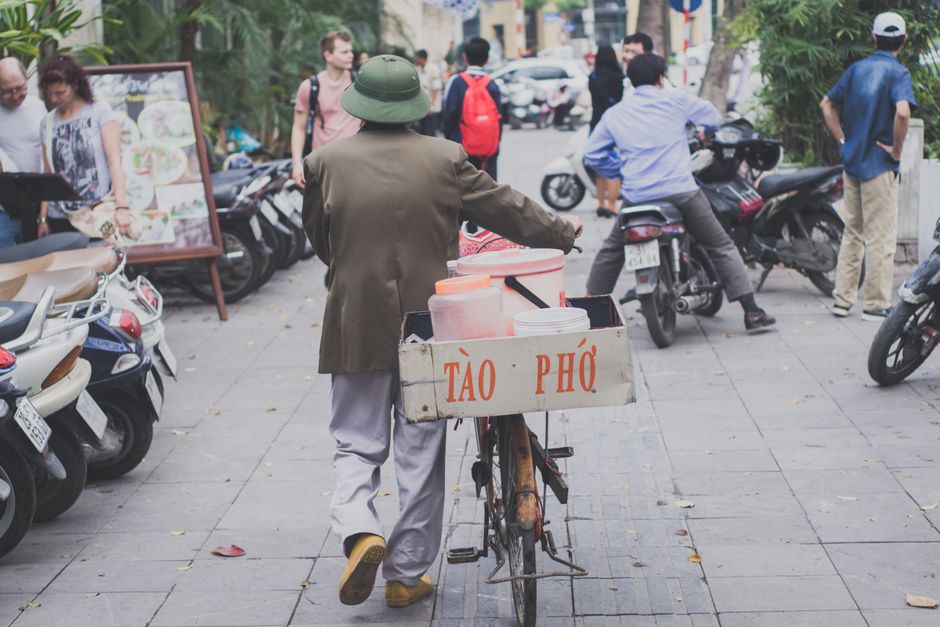 Man pushes bicycle food cart on bustling city street with people and motorcycles.